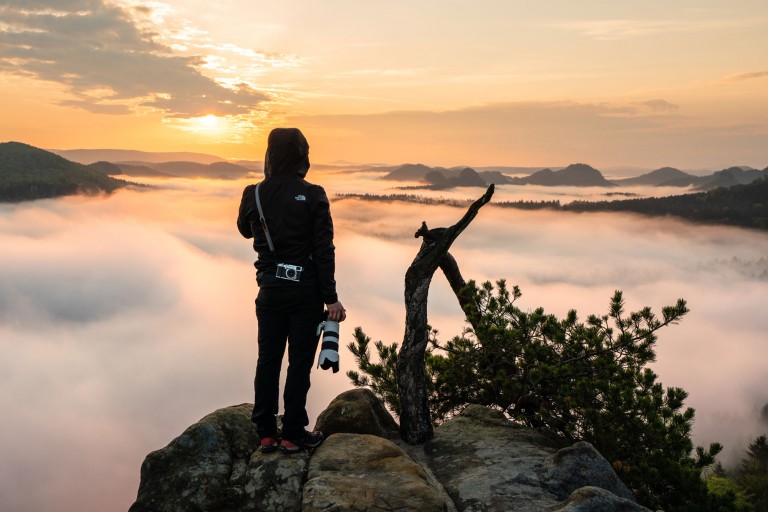 Eine Person in schwarzer Jacke und Hose steht auf einer Felsklippe, hält eine Kamera mit großem Objektiv in der Hand und blickt bei Sonnenaufgang auf ein nebliges Tal. Die Szene umfasst sanfte Hügel und einen teilweise bewölkten Himmel, mit etwas Grün und einem Ast im Vordergrund.