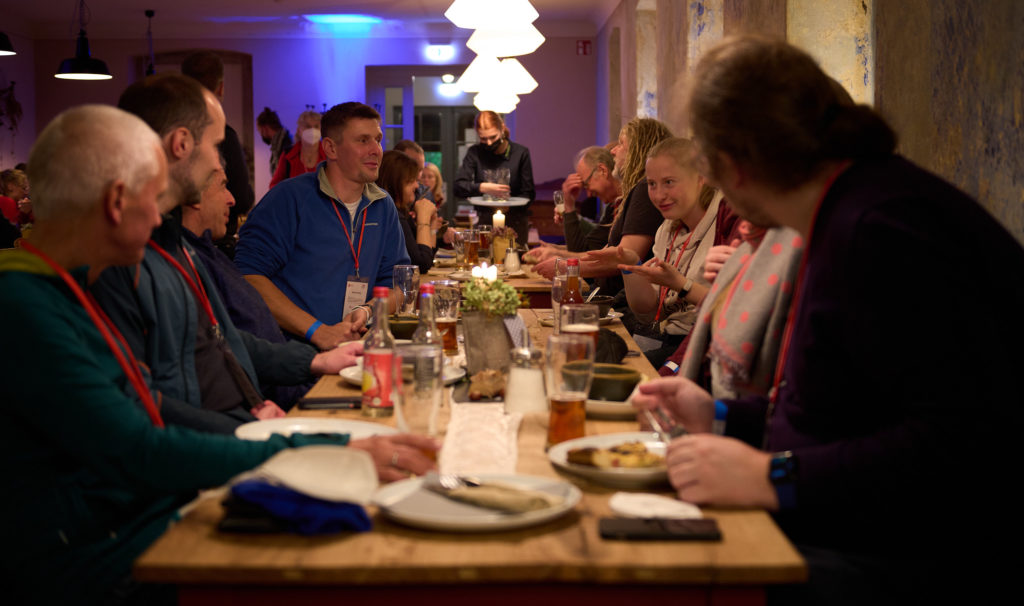 Eine Gruppe von Menschen sitzt um einen langen Holztisch in einem warm beleuchteten Restaurant. Sie unterhalten sich, essen und trinken Bier. Im Hintergrund sind andere Gäste und festliche Dekoration zu sehen, was für eine lebhafte und gemütliche Atmosphäre sorgt.