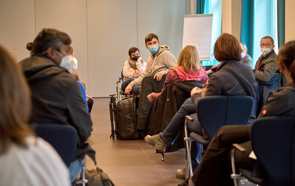 Eine Gruppe von Menschen mit Gesichtsmasken sitzt in einem Konferenzraum im Halbkreis. Eine Person in der Mitte dreht sich auf ihrem Stuhl um und unterhält sich mit anderen. Im Hintergrund ist ein Flipchart zu sehen.