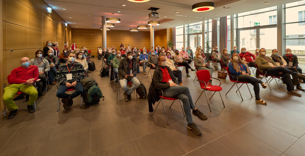 Eine Gruppe von Menschen sitzt auf roten Stühlen in einem Konferenzraum. Alle Teilnehmer tragen Mund-Nasen-Schutz. Der Raum hat Holzwände, große Fenster und Deckenleuchten mit roten Akzenten. Einige Menschen haben Laptops oder Notebooks auf dem Schoß und blicken aufmerksam nach vorne.