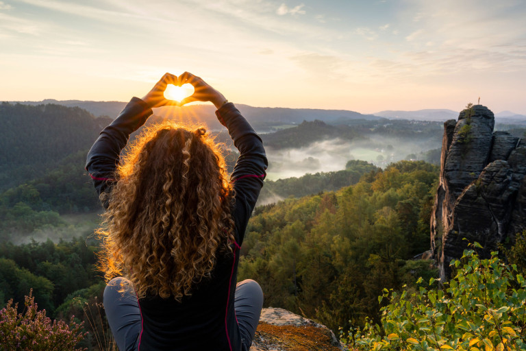 Eine Person mit lockigem Haar sitzt auf einem Felsvorsprung und blickt auf eine malerische, neblige Landschaft bei Sonnenaufgang. Sie hält ihre Hände in Form eines Herzens hoch und umrahmt damit die aufgehende Sonne, mit üppigen grünen Bäumen, Hügeln und entfernten Klippen im Hintergrund.