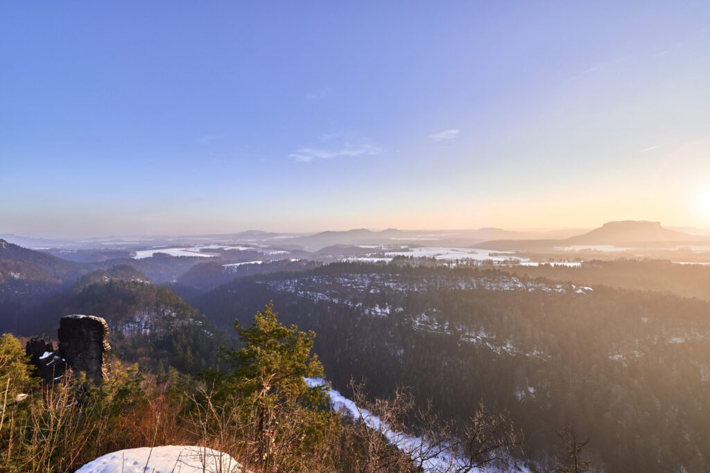 Ein Panoramablick auf eine ruhige Landschaft bei Sonnenaufgang. Die Szene zeigt sanfte Hügel, die mit immergrünen und laubabwerfenden Bäumen bedeckt sind, Schneeflecken und eine entfernte Bergkette, die im sanften goldenen Licht der Morgensonne getaucht ist.