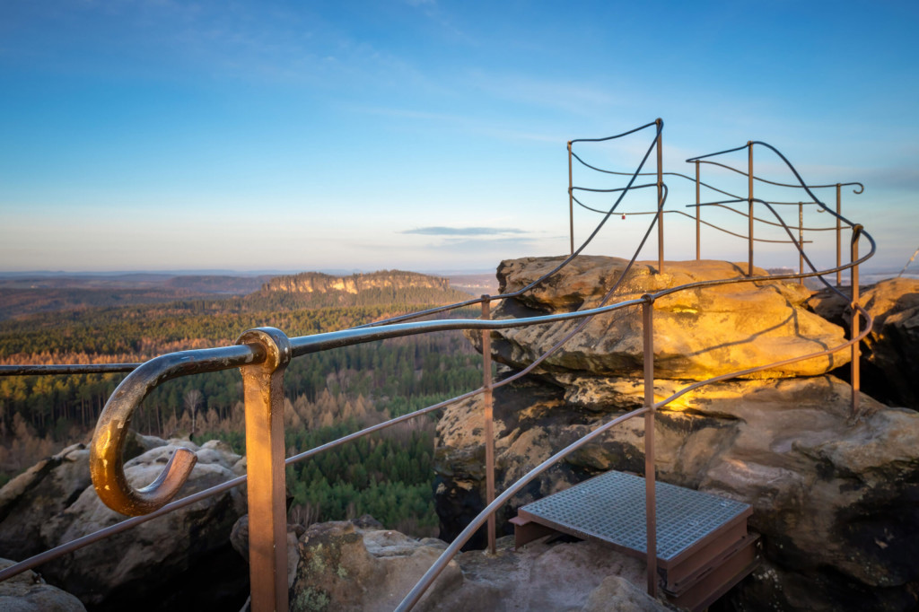 Ein Aussichtspunkt mit Metallgeländer auf einem Felsvorsprung bietet bei Sonnenuntergang einen Blick auf eine weite Landschaft aus Wäldern und Hügeln unter einem klaren blauen Himmel. Die Felsen und Geländer werden vom Licht der goldenen Stunde beleuchtet.