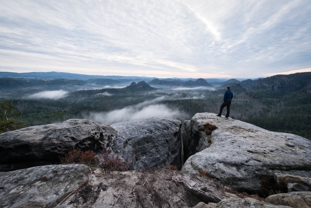 Eine Person steht am Rand einer Felsklippe und überblickt eine weite Landschaft mit bewaldeten Hügeln und Tälern. Nebel oder tief hängende Wolken schweben über den Bäumen, und der Himmel ist mit einer Schicht vereinzelter Wolken bedeckt, was eine dramatische und heitere Szene schafft.