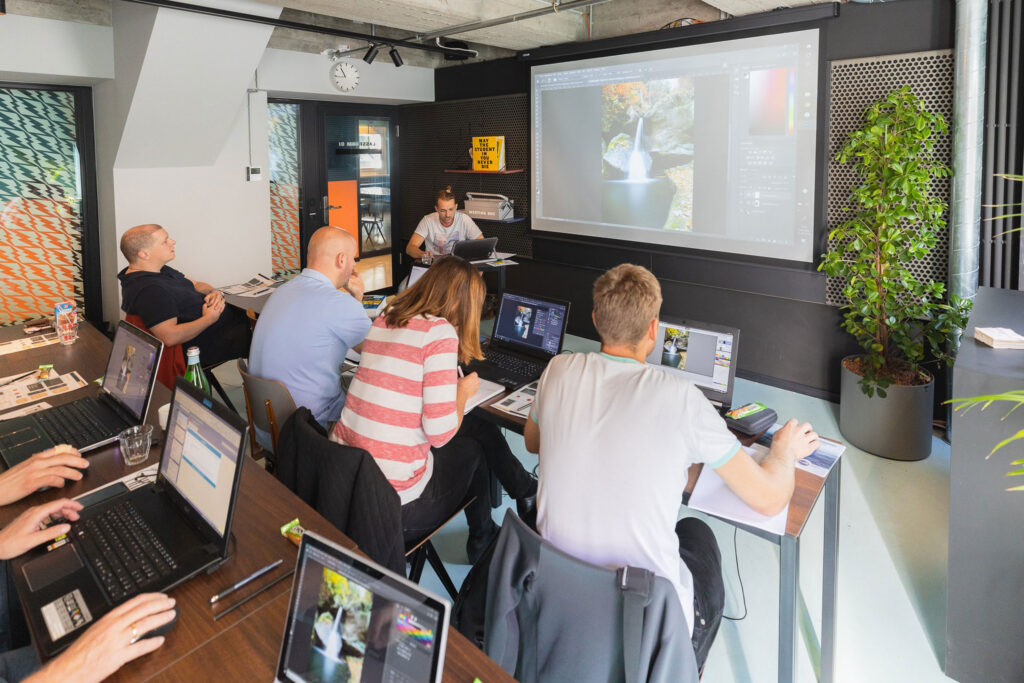 Ein Klassenzimmer mit Menschen, die an Laptops arbeiten und sich auf Bildbearbeitungssoftware konzentrieren. Ein Projektor zeigt ein Bild, das bearbeitet wird. Ein Mann steht davor und gibt Anweisungen. Eine große Pflanze und gemusterte Wandpaneele schmücken den Raum.