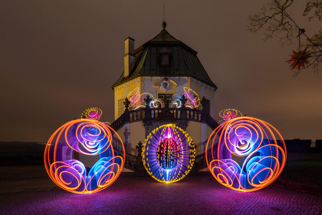 Ein historisches Gebäude wird nachts von lebendigen, wirbelnden Mustern aus orangefarbener, blauer, rosa und violetter Lichtkunst beleuchtet, die auf beiden Seiten des Eingangs große kreisförmige Formen erzeugen.