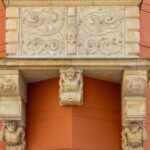 Verschnörkelter Steinbalkon mit kunstvollen Schnitzereien, getragen von menschlichen Figuren und Löwenköpfen, vor einem orangefarbenen Gebäude mit weiß gerahmten Fenstern.