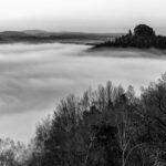 Ein Schwarz-Weiß-Foto von einer nebligen Landschaft mit dichtem Nebel, der die Täler bedeckt. Baumbewachsene Hügel erheben sich über den Nebel, und im Vordergrund sind kahle Äste zu sehen. Die Szene wirkt ruhig und geheimnisvoll.