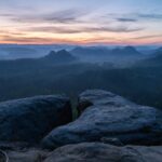 Blick von felsigen Klippen über einen nebelverhangenen Wald und sanfte Hügel bei Sonnenaufgang, mit sanften orangefarbenen und blauen Tönen am Himmel und Schichten von fernen Bergen, die am Horizont verschwinden.