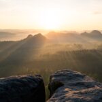 Sonnenaufgang über einer nebligen Berglandschaft mit sanftem Sonnenlicht, das durch die Wolken und bewaldeten Hügel dringt. Zwei große Felsen im Vordergrund überblicken die malerische Aussicht.