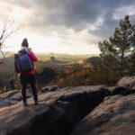 Zwei Wanderer mit Rucksäcken stehen auf einem großen felsigen Gelände mit Blick auf ein malerisches Tal mit herbstlichen Bäumen, fernen Hügeln und dramatischen Wolken, die vom goldenen Sonnenlicht beleuchtet werden.