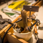 Eine weiße Tasse hält verschiedene Metall- und Holzwerkzeuge, darunter stempelartige Werkzeuge mit gemusterten Enden, die auf einer unübersichtlichen Werkbank mit Papieren und anderen unscharfen Gegenständen im Hintergrund liegen.