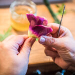 Hände, die mit einem dünnen Metallwerkzeug eine Blume aus violetten und gelben Blütenblättern formen, mit einem Glas mit gelber Flüssigkeit und grünen Blättern auf einem Holztisch im Hintergrund.