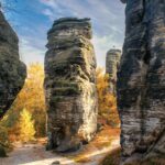 Hohe, verwitterte Felsformationen erheben sich zwischen herbstlichen Bäumen mit goldenen Blättern unter einem teilweise bewölkten Himmel und schaffen eine dramatische, natürliche Landschaft. Das Sonnenlicht wirft Schatten auf den sandigen Boden und hebt die Textur der Felsen hervor.
