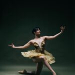Eine Ballerina in einem goldenen Tutu posiert anmutig auf einem Knie, die Arme elegant ausgestreckt, vor einem dunklen Hintergrund.