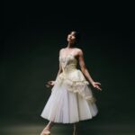 Eine Ballerina in einem weiß-goldenen Tutu posiert anmutig auf Spitzen vor einem dunklen Hintergrund, die Arme leicht ausgestreckt und den Kopf nach oben geneigt.