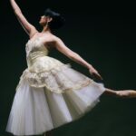 Eine Balletttänzerin in einem cremefarbenen Tutu nimmt vor einem dunklen Hintergrund eine elegante Pose auf Spitzen ein, wobei sie einen Arm nach oben und den anderen zur Seite ausgestreckt hält.