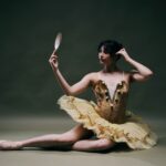 Vor einem dunklen, neutralen Hintergrund sitzt eine Ballerina in einem gold-gelben Tutu auf dem Boden, ein Bein ausgestreckt; in der einen Hand hält sie einen kleinen Spiegel, mit der anderen richtet sie sich die Haare.