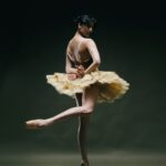 Eine Ballerina in einem goldenen Tutu und Ballettschuhen posiert anmutig auf Spitzen vor einem dunklen, schlichten Hintergrund, wobei sie ein Bein hinter dem anderen kreuzt und die Arme elegant ausstreckt.