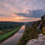 Ein malerischer Fluss schlängelt sich zwischen bewaldeten Hügeln und felsigen Klippen unter einem farbenprächtigen, bewölkten Abendhimmel hindurch, während die Bäume ihr Herbstlaub zur Schau stellen und vereinzelt Häuser in der Landschaft zu erkennen sind.