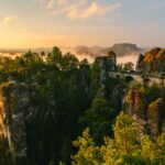 Eine Steinbrücke überspannt bei Sonnenaufgang dramatische, von Grün bewachsene Felsformationen, vor dem Hintergrund von Nebel und sanftem goldenem Licht, umgeben von Wald und fernen Bergen.