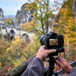 Eine Person richtet eine Kamera auf einem Stativ aus und fokussiert dabei eine malerische Steinbrücke, die von felsigen Klippen und herbstlichen Bäumen im Hintergrund umgeben ist. Auf dem Bildschirm der Kamera ist dieselbe malerische Landschaft zu sehen.