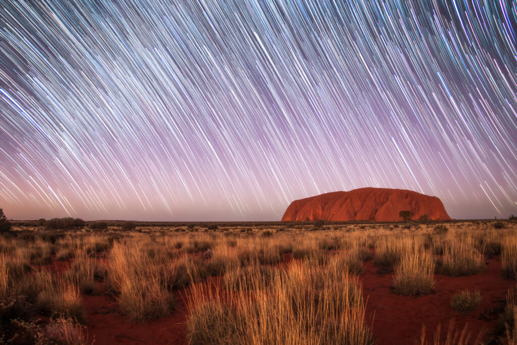 Der Uluru ragt aus einer rötlichen Wüstenlandschaft unter einem Himmel voller Sternspuren empor, mit Gräsern im Vordergrund und weißen und blauen Lichtstreifen, die sich bogenförmig über den Nachthimmel ziehen.