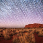 Der Uluru ragt aus einer rötlichen Wüstenlandschaft unter einem Himmel voller Sternspuren empor, mit Gräsern im Vordergrund und weißen und blauen Lichtstreifen, die sich bogenförmig über den Nachthimmel ziehen.