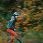 Eine Person mit Helm, grüner Jacke und roter Hose fährt im Freien auf einem Einrad, wobei das verschwommene Herbstlaub im Hintergrund einen Eindruck von Bewegung vermittelt.
