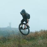 Eine Person, die einen Helm und Schutzkleidung trägt, führt an einem nebligen Tag auf einem Mountainbike einen Sprung über grasbewachsenes Gelände aus; im Hintergrund sind Hügel und ein Radar-Turm in der Ferne zu sehen.