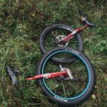Zwei Einräder mit großen Reifen liegen auf grünem Gras und inmitten von Pflanzen, daneben ein Paar schwarze Fahrradschuhe; die Szene lässt auf eine Pause während einer Offroad-Tour mit dem Einrad schließen.
