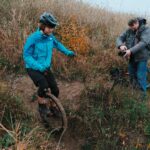Eine Person in einer blauen Jacke und mit Helm fährt auf einem Einrad einen Feldweg entlang, während eine andere Person mit einer Kamera in der Nähe kniet und die Szene inmitten von hohem Gras und Laub fotografiert.